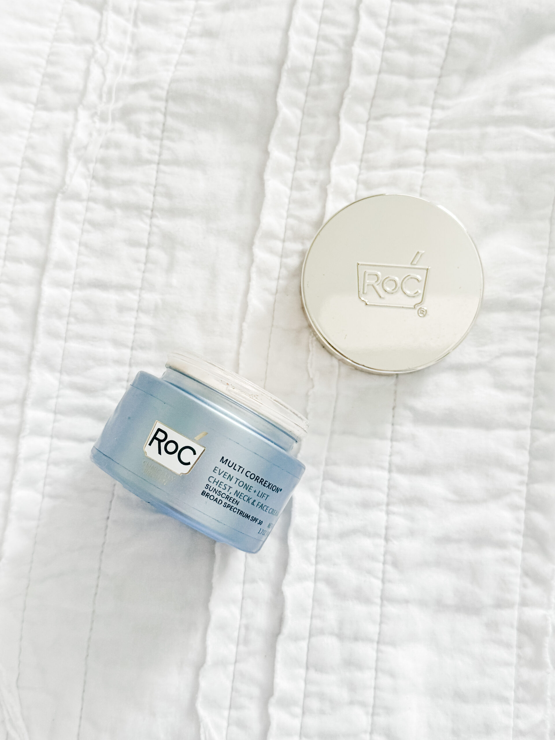 Top view of an open RoC Multi Correxion Cream jar revealing peach-toned cream inside, with the silver lid placed beside it on a white quilt.